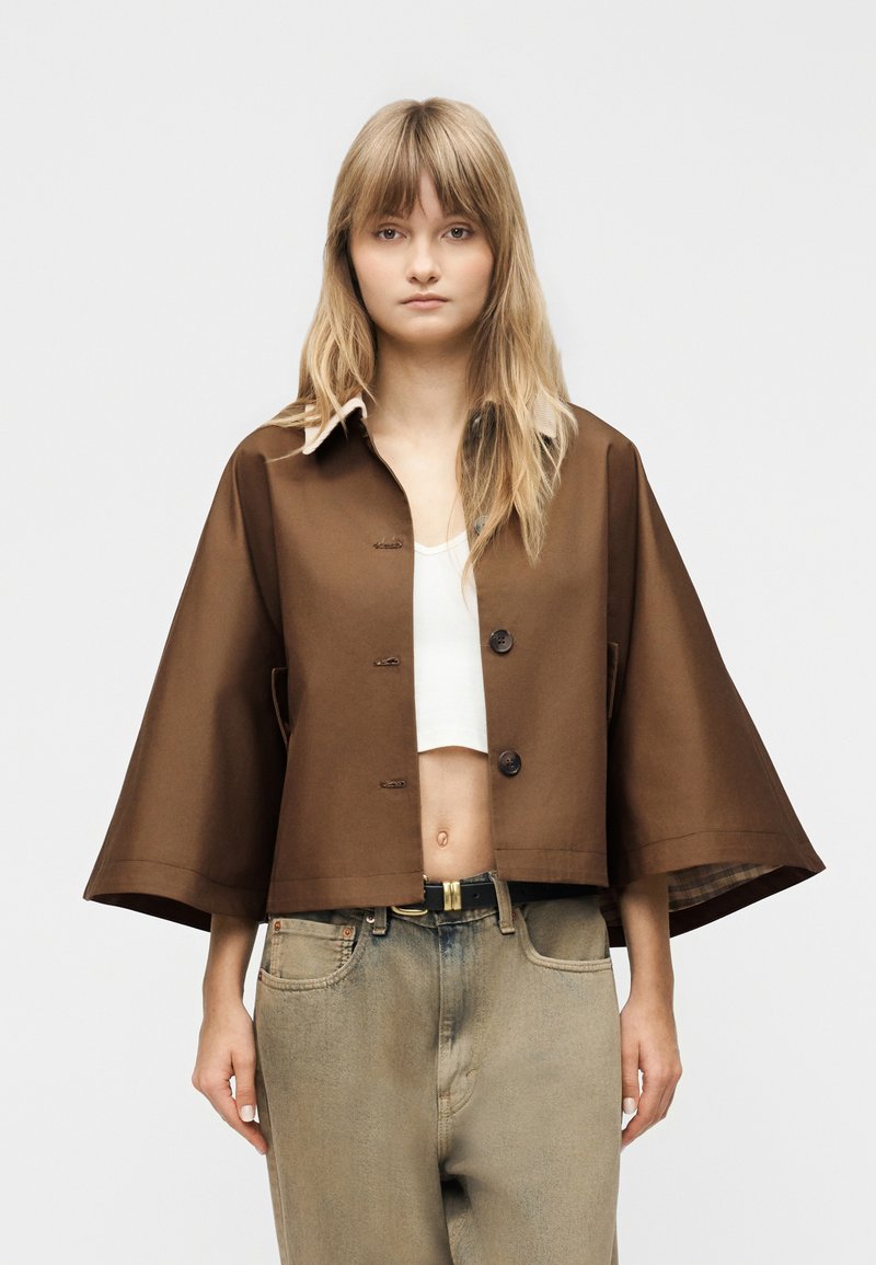 Ung kvinde iført en brun cropped jakke med brede ærmer, hvid cropped top og højtaljede falmede jeans mod en ensfarvet baggrund.