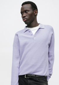 Suéter texturizado de color púrpura claro con cuello tipo polo, tres botones y mangas largas, llevado sobre una camisa blanca. Cinturón negro con hebilla plateada.