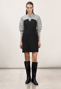 Vestido de tirantes negro con escote en V y dos bolsillos frontales, superpuesto a una blusa de rayas en blanco y negro. El modelo lleva botas altas negras.