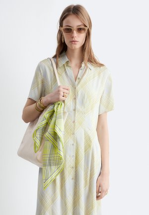 Donna che indossa un vestito camicia a quadri giallo chiaro, occhiali da sole beige, gioielli in oro, con una borsa beige e una sciarpa verde fantasia.