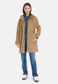 Camel-Wollmantel mit Kapuze, vorderer Druckknopfverschluss und großen Taschen. Kombiniert mit einem Jeanshemd, blauen Jeans und weißen Turnschuhen.