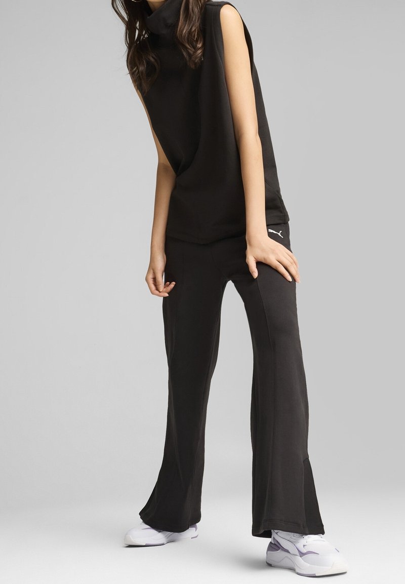 Femme portant un haut sans manches noir et un pantalon avec des fentes latérales, assortis à des baskets blanches et violettes sur un fond gris.