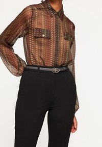 Blusa trasparente e a fantasia in toni terreni di bordeaux e verde, con apertura frontale a bottoni, colletto e due tasche sul petto. Abbinata a pantaloni neri.