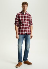 Camicia a scacchi rossa e bianca con maniche arrotolate, abbinata a jeans blu strappati e scarpe marroni. Design semplice e stile casual.