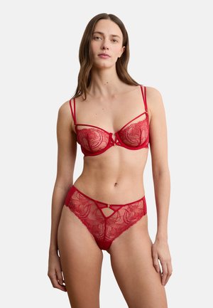 Kobieta w czerwonym koronkowym biustonoszu i dopasowanych przezroczystych koronkowych majtkach, stojąca na tle jednolitego białego tła.