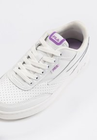 Baskets blanches Fila avec bout perforé, lacets blancs, logo Fila violet sur la languette et le côté, et doublure intérieure matelassée visible.