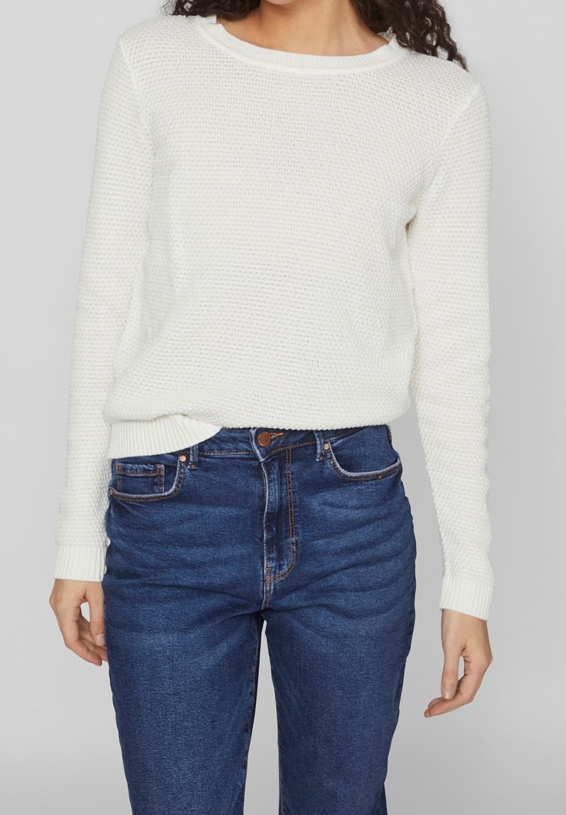 Pull en maille blanche avec motif texturé, col rond, poignets et ourlet côtelés, associé à un jean en denim bleu taille haute.