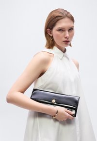 Νεαρή γυναίκα με κοντά μαλλιά, φορώντας λευκό αμάνικο μπλουζάκι, κρατά μαύρο γυαλιστερό τσαντάκι φάκελο με χρυσό κούμπωμα σε απλό φόντο.