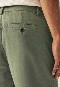 Pantalon en coton vert avec une poche arrière dotée d'un bouton noir, texture lisse et coupe ajustée, présenté de la taille à la hanche.