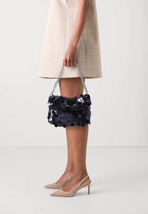Femme en robe beige et talons, tenant un sac à main noir en sequins avec une bandoulière chaîne argentée, sur un fond blanc uni.