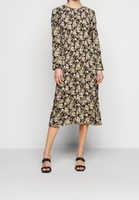 Långärmad blommig klänning i svart med mönster i krämfärg och beige. Har en rund halsringning och en flödande siluett. Matchas med svarta sandaler.