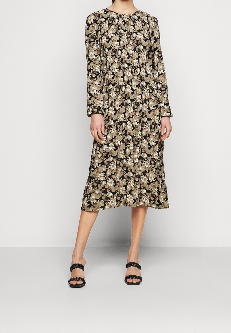 Långärmad blommig klänning i svart med mönster i krämfärg och beige. Har en rund halsringning och en flödande siluett. Matchas med svarta sandaler.