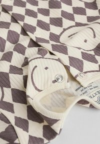 Cremfarbener Onesie mit kariertem Muster in Dunkelbraun. Enthält Druckknöpfe und gerippte Textur, mit einem sichtbaren Etikett im Inneren.