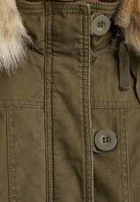 Olivově zelená parka s límcem z umělé kožešiny, vybavená dvěma knoflíky, vertikálními švy a našitými kapsami. Texturovaná látka s odolným vzhledem.