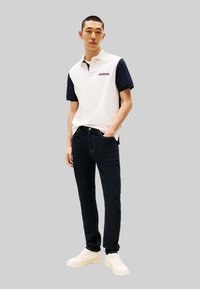 Weißes und marineblaues Poloshirt mit Logo, kombiniert mit dunklen Jeans und weißen Turnschuhen. Das Outfit verfügt über ein Design mit Kragen und kurze Ärmel.