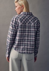 Plaid shirt in purple, gray, and black, with a curved hem and buttoned cuffs. Paired with light gray jeans. Textured fabric with visible seams.