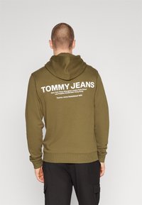 Muž v olivově zelené mikině s kapucí s nápisem "TOMMY JEANS" a mezinárodními názvy měst vytištěnými bílou barvou na zádech, doplněné černými kalhotami.