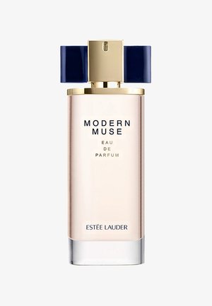 Heldere glazen fles met een verloop van helder naar lichtroze, gouden accenten, marineblauw deksel en een "MODERN MUSE" label aan de voorkant.