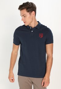 Polo din bumbac de culoare albastru marin, cu un guler cu trei nasturi și un logo brodat roșu pe pieptul stâng. Textură netedă.