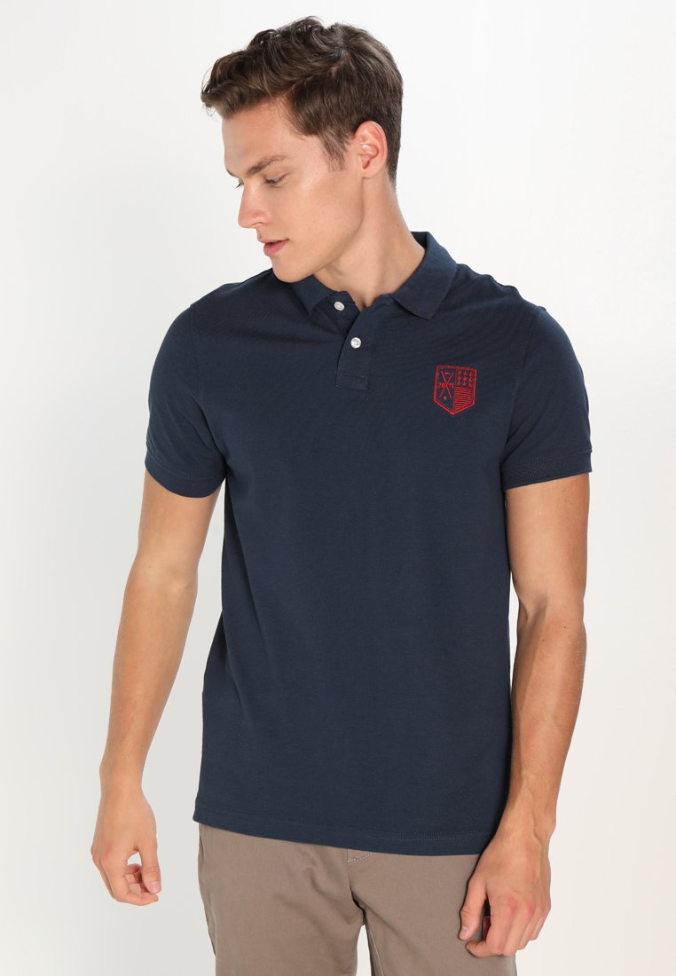 Polo din bumbac de culoare albastru marin, cu un guler cu trei nasturi și un logo brodat roșu pe pieptul stâng. Textură netedă.
