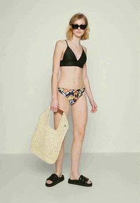 Top de bikini triangular preto, parte de baixo de bikini com padrão floral, bolsa de palha em tom bege, sandálias slides pretas, modelo com cabelo curto e óculos de sol.