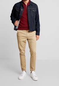 Giacca in denim scuro, felpa con zip bordeaux, chinos beige e sneakers bianche. La giacca presenta due tasche sul petto e un taglio classico.
