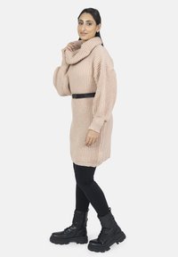 Vestido de suéter beige de punto con cuello cisne y mangas abullonadas, ceñido en la cintura con un cinturón negro. Combinado con botas de combate negras.