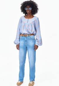 Kobieta o kręconych włosach i dużych okularach przeciwsłonecznych, ubrana w jasnoniebieską kratowaną bluzkę, brązowy pasek, jasnoniebieskie dżinsy i beżowe trampki na białym tle.