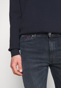 Suéter de marina con puños acanalados, combinado con jeans de mezclilla oscuros que presentan un sutil desgastado y un acento rojo en el bolsillo. Mano posicionada en el lado.