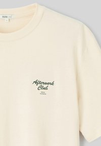 T-shirt couleur crème en tissu doux, orné d'un petit graphique vert avec l'inscription "Afterwork Club" et "partagez des moments" en dessous.