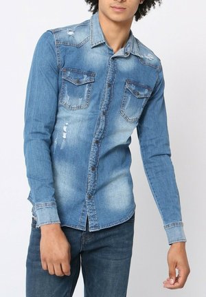 Chemise en denim bleu clair avec des manches longues, fermeture à boutons, poches poitrine et détails abîmés. Les sections décolorées accentuent la texture.