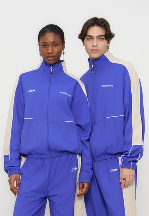 Két modell passzoló kék és bézs melegítőfelsőben, melyeken a "YOURTURN" logó látható, állnak egymás mellett egy egyszerű háttér előtt.