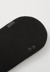 Calcetín negro hecho de una tela suave y lisa. Presenta el texto "BOSS" en gris y detalles de tamaño en una fuente pequeña cerca del borde. Diseño de punta redondeada.