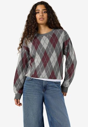 Femme aux longs cheveux bouclés portant un pull à losanges gris et bordeaux et un jean bleu, debout devant un fond uni.