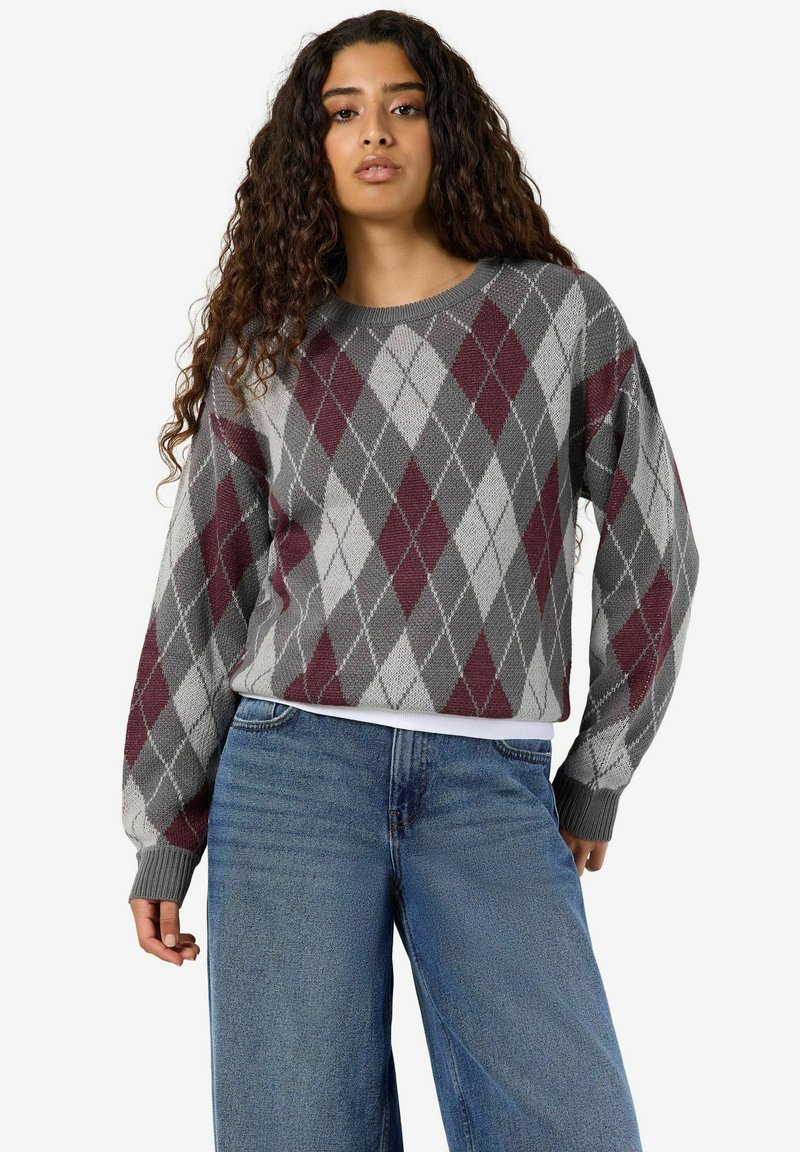 Femme aux longs cheveux bouclés portant un pull à losanges gris et bordeaux et un jean bleu, debout devant un fond uni.