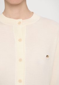 Ljusbeige cardigan med rund hals, fem knappar framtill och en liten broderad design på vänster bröst. Mjuk textur.