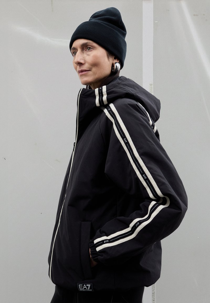 Črna jopa s kapuco z belimi črtami ob rokavih, z zadrgo za zapiranje in elastičnimi manšetami. Nošeno s temno kapa.