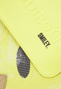 Żółta torba z syntetycznej skóry z teksturowaną powierzchnią, wytłoczonym napisem "designed" oraz czarnym logo "SMILEY" na tkanym tle.