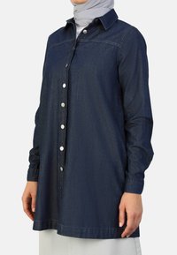 Donkerblauwe denim overhemd met een knoopsluiting aan de voorkant, lange mouwen met manchetten en een klassieke kraag. Gladde textuur met minimale stiksels.