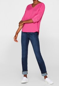 Maglione fucsia a V con maniche a tre quarti e dettagli con bottoni, abbinato a jeans scuri arrotolati e sneakers bianche.