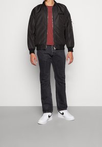 Chaqueta bomber negra con cremallera, camiseta burdeos, jeans oscuros y zapatillas blancas con detalles negros. Tejido suave y diseño ajustado.