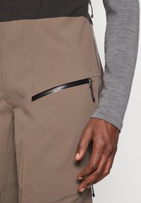 Desna ruka pored smešnog donjeg dela s uglastim crnim džepom s patent-zatvaračem, uparena s sivom majicom dugih rukava; klizači patent-zatvarača vidljivi na odjeći.