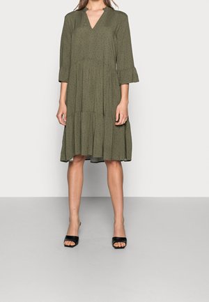 Woman wearing an olive green, knee-length, tiered dress with small black dots, three-quarter sleeves, and black open-toe mule heels.