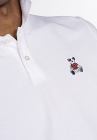 Camisa polo blanca con cuello, que presenta un pequeño oso de peluche bordado con una camiseta roja en el lado izquierdo.