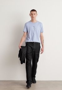 Modelo que lleva una camiseta de algodón azul claro con un logo, pantalones negros texturizados y zapatillas negras, sosteniendo una chaqueta negra.