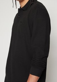 Svart långärmad polo-shirt i ett strukturerat stickat material med krage, och en subtil broderad logotyp på bröstet.
