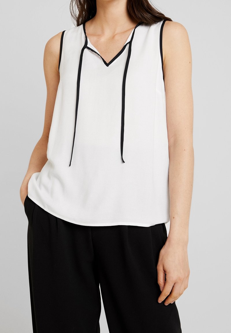Femme portant un chemisier blanc sans manches avec un bord noir et un pantalon noir, debout avec une main dans la poche sur un fond uni.
