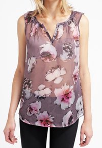 Top trasparente viola con un motivo floreale in rosa e bianco. Presenta un colletto arricciato e una vestibilità comoda. Abbinato a pantaloni neri.