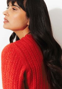 Pull en tricot rouge avec un motif de câble vertical, encolure ronde et bords côtelés. Cheveux bruns coiffés en vagues lâches. Boucles d'oreilles en or en forme de hoop.