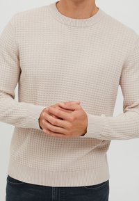 Pull beige clair en maille avec un motif texturé, col rond, poignets et ourlet côtelés, porté par une personne les mains jointes.